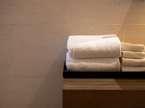 White clean folded towels in the black tray on wooden table white marble tiles wall background with copy space. Towel set reserved for the guest in hotel room, with warm light.