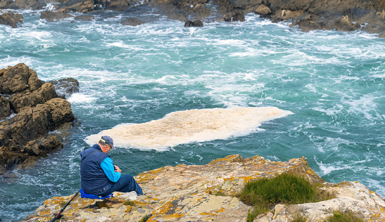 Senior and retired man in blue fisherman's cap and vest preparing his fishing rod and hooks sitting on the cliff of Rinlo fishing village with rough sea waves and foam.