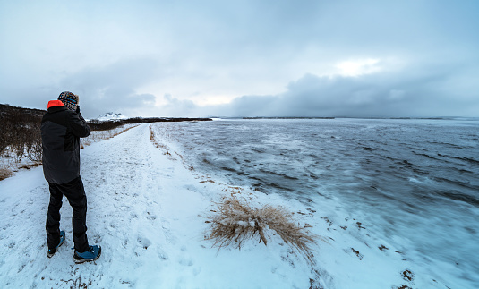 Man standing protected by the polar cold with jacket, hat and boots, taking photos of a completely snow covered road and landscape and a frozen river in Iceland