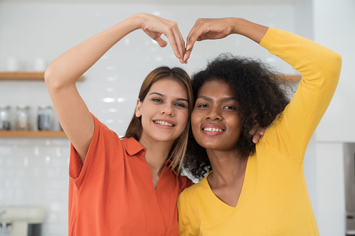 Happy two young women embracing and showing heart symbol with their hands at home. Two young diverse lesbian women spending time together at home. Diversity, LGBT and gender identity concept