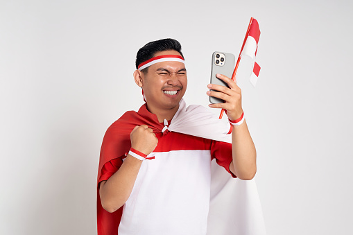 Excited handsome young Asian man in casual white t-shirt holding smartphone and Indonesian flag isolated over white background. Celebrate Indonesia independence day on 17 August