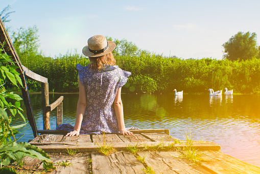 Beautiful girl is sitting on the wooden pier. Rustic and natural photo outdoors. Summer river. Relax and travel concept.