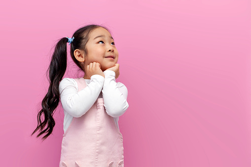 little pensive asian girl in pink sundress looks away and dreams on pink isolated background, korean preschool child thinks and is puzzled by question