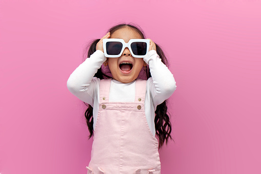 little shocked preschool girl in white sunglasses and pink sundress screams and is surprised on pink background, the child looks into glasses with her mouth open
