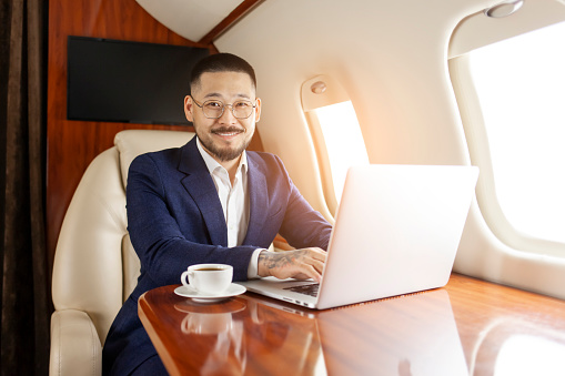 successful asian businessman in suit and glasses sits in private jet and types on laptop, korean entrepreneur in business clothes flies in airplane, luxury lifestyle