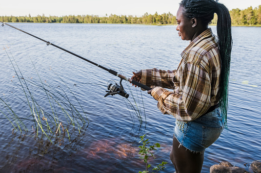 Woman stands and reels in the fishing line during a fishing trip in one of the lakes in Sweden on a sunny summer day.