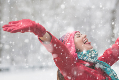 Happy woman in red jacket and warm knitted scarf catches snowflakes in winter forest on snowy day, sunlight
