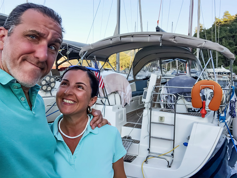 Loving middle aged caucasian male adult with blue eyes looking at camera and taking a selfie with his toothy smiling middle eastern woman in front of a sailing boat on a summer day.