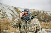 Soldier is standing in the camouflage uniform and checkered keffiyeh shemagh bandana. Binocular in hands. Terrorist is outdoors in the abandoned deserted place. War concept.