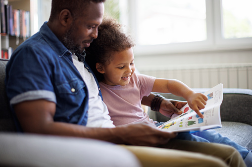 Happy African American father and adorable mixed race daughter are reading a book and smiling while spending time together at home. Children education and development concept.