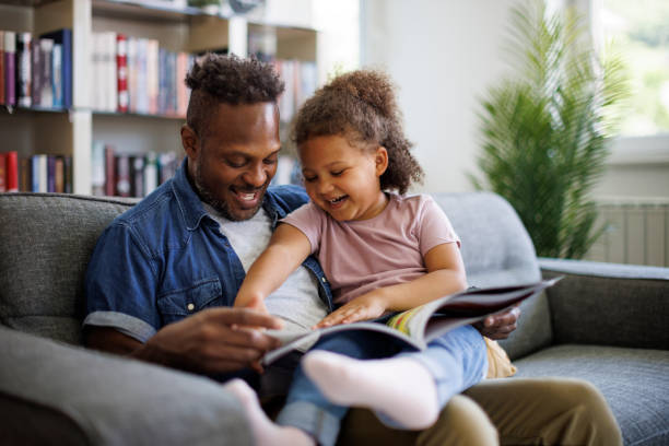 행복한 아프리카계 미국인 아버지와 사랑스러운 혼혈 딸이 집에서 함께 시간을 보내며 책을 읽고 웃고 있다. 어린이 교육 및 발달 개념 스톡 사진