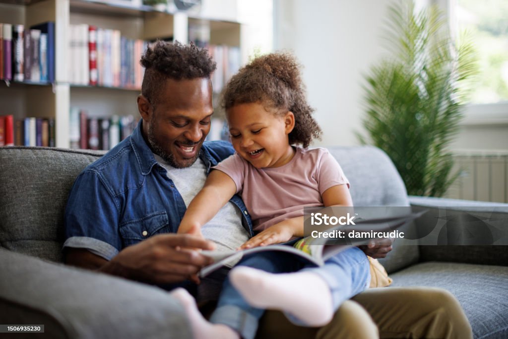 Happy African American father and adorable mixed race daughter are reading a book and smiling while spending time together at home. Children education and development concept Reading Stock Photo