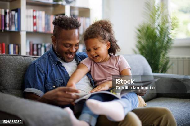 Happy African American Father And Adorable Mixed Race Daughter Are Reading A Book And Smiling While Spending Time Together At Home Children Education And Development Concept Stock Photo - Download Image Now