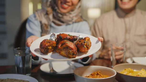 Happy Asia muslim wife and husband Ramadan dinner together show fried chicken indian food plate in dining room at home to camera. Family catering celebration end of Eid al-Fitr togetherness home. stock photo