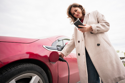 Woman with smartphone charging red electric car