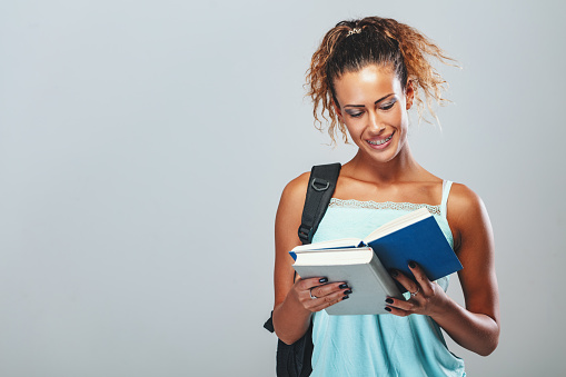 Beautiful smiling teenage girl with school bag is reading a book.