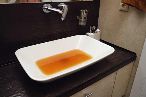 The rusty water gushes out of the faucet, making the sink wet and dirty. The unclean water flowing from the rusty faucet in the bathroom is unfit for use.