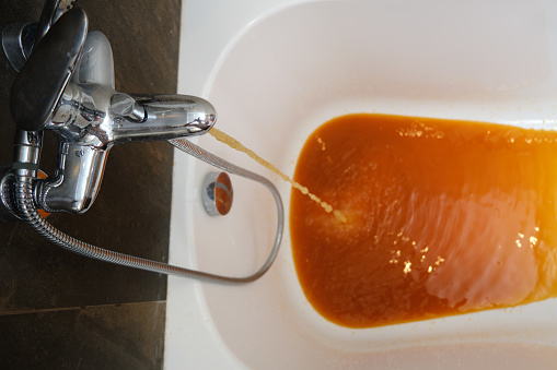 The faucet in the bathroom releases a stream of brown liquid. The flowing water from the faucet turns orange due to rust.