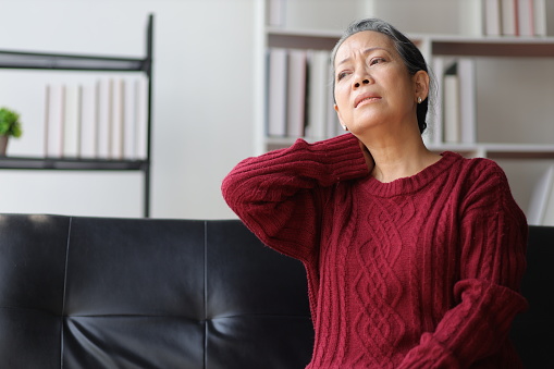 Senior woman sitting on the sofa at home is sick and has neck pain.