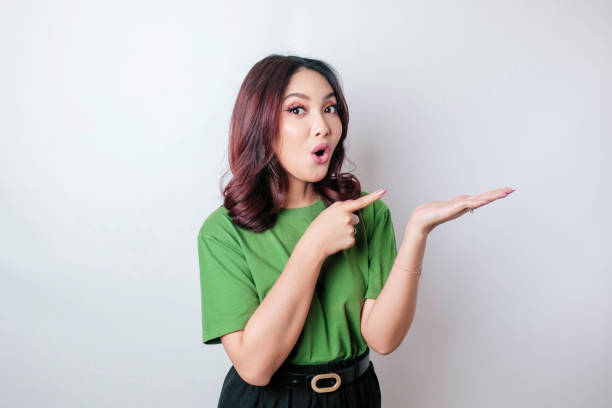shocked asian woman wearing green t-shirt pointing at the copy space beside her, isolated by white background - copy space left imagens e fotografias de stock