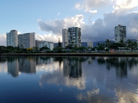 Waterfront with iconic cityscape in the backdrop. The Ala Wai Canal is a man-made waterway that runs through the city of Honolulu on the island of Oahu. It is lined with condos, golf courses, and coconut trees. In the distance, the mountains of the Ko'olau Range can be seen. This panoramic photo shows the beauty and tranquility of this popular tourist destination.