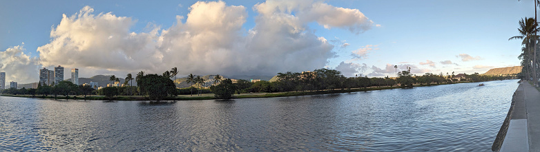 The Ala Wai Canal is a man-made waterway that runs through the city of Honolulu on the island of Oahu. It is lined with condos, golf courses, and coconut trees. In the distance, the mountains of the Ko'olau Range can be seen. This panoramic photo shows the beauty and tranquility of this popular tourist destination.