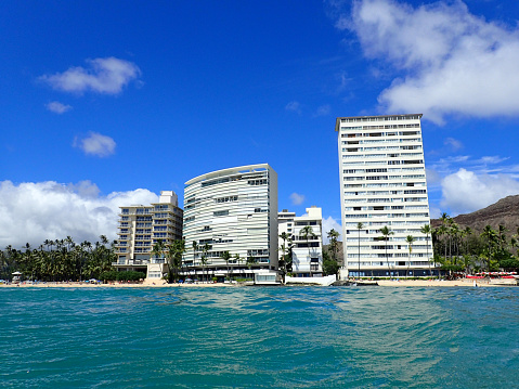 A beautiful view of the wavy water on the ocean off Kaimana Beach, a popular spot for swimming and surfing on the south shore of Oahu, Hawaii. The skyline of Waikiki, with its hotels and condos, can be seen in the background. The image captures the contrast between the natural beauty of the sea and the urban development of the city.