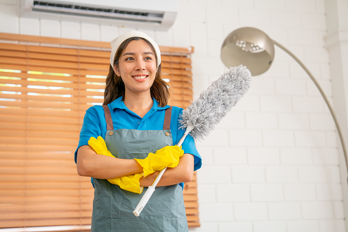 Portrait of Asian housekeeper or housemaid stand with arm-crossed with smiling and look forward also hold  feather duster in living room with day light.