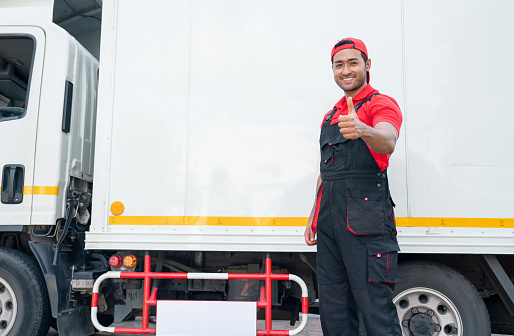 Portrait of Asian delivery man stand beside of truck and show thumbs up also look at camera with smiling after finish to send the product to customer house.