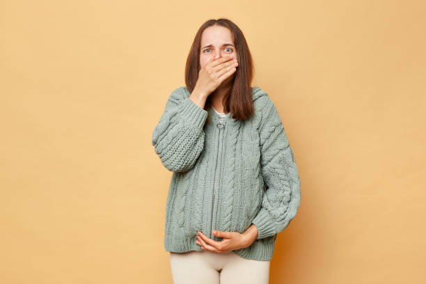 istock Unhealthy pregnant woman wearing knitted warm sweater standing isolated over beige background felling nausea covering mouth with palm bad feeling sick future mother. 1507553473