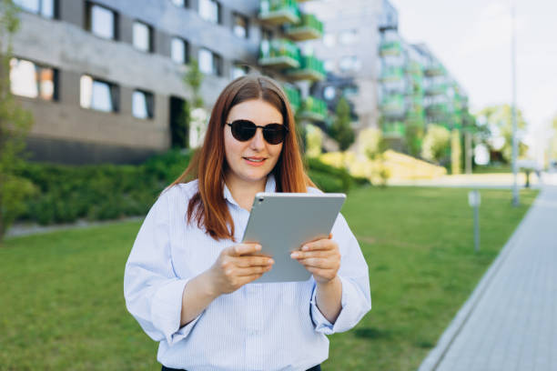 istock Redhead woman executive smiling confident using touchpad on urban background. Happy young women in a blue shirt and sunglasses walks down the street. Successful business. 1507611604