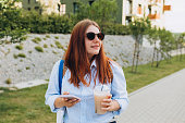 Outdoor lifestyle fashion portrait of woman posing on the street drinking smoothie or ice coffee. Close up of woman holding eco or reusable shopping bag and using smartphone