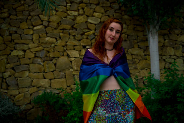 mujer sonriente de pie contra un muro de piedra envuelto en la bandera del arco iris - homosexual gay pride business rainbow fotografías e imágenes de stock