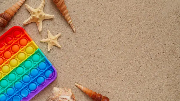 Photo of Beach summer vacation banner concept with trendy bright rainbow silicone stress relief toy.Colorful pop it toy,seashells and starfishes lie sand,top view,flat lay,copy space.Fashionable fidgeting game