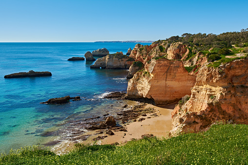 Solitary beach close to Alvor village in Portugal. There are lots of limestone rocks in the water and green grass in the foreground.
