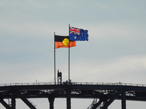 The Australian and Australian Aboriginal flags fly on the Sydney Harbour Bridge on a windy and sunny afternoon in winter.  At the base of the flags is the silhouette of a group of people climbing the bridge. This image was taken from Mrs Macquarie's Chair on 1 July 2023.