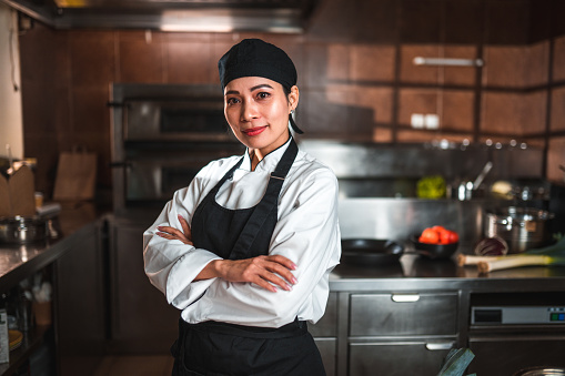 Front portrait of a beautiful chinese chef standing in the kitchen. She is wearing a white uniform and has her arms crossed.