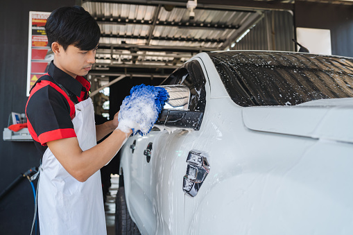 Asian male employee hand cleaning a car by rubbing a silver cloth over a white car.