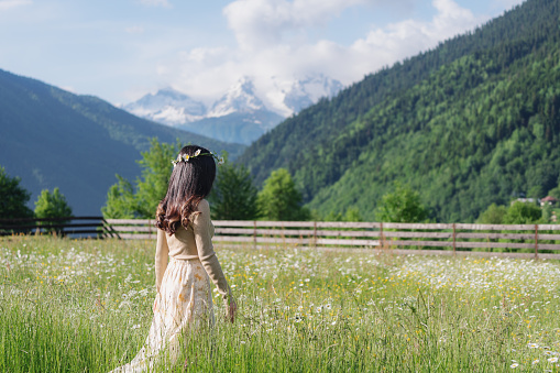 Woman with flowers crown, walking in field of flowers, with natural landscape of trees and mountain