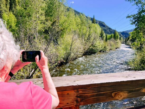 Senior Tourist Woman Taking a Photo of a River in Colorado in the Late Spring Cheerful Senior Woman Using a Smartphone to Take a Photo of Tenmile Creek in Frisco, Colorado While Traveling in the Mountains tenmile range stock pictures, royalty-free photos & images