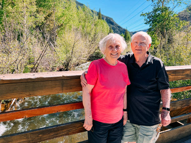 Cheerful Senior Couple Standing on a Bridge Together in Colorado While Traveling in the Mountains Cheerful Senior Couple Standing on a Bridge Together at Tenmile Creek in Frisco, Colorado near Breckenridge While Traveling in the Mountains in the Summer tenmile range stock pictures, royalty-free photos & images