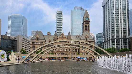 view to the Old City Hall of Toronto, modern buildings behind, with cloudy sky. the Arch and Fountain at Nathan Phillips Square in front.