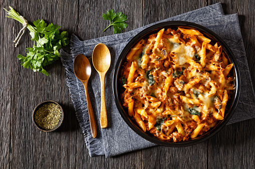 Ground Chicken Pasta Bake with onion, mushrooms, spinach, tomato sauce and mozzarella cheese in baking dish on dark wood table with spoons, horizontal view from above, flat lay, copy space