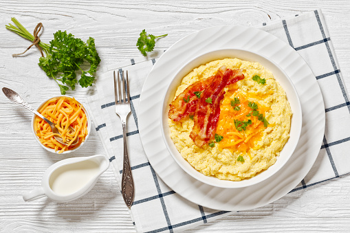 Cheddar Cheese Grits Casserole with fried bacon slices in white bowl on white wooden table with ingredients, horizontal view from above, flat lay