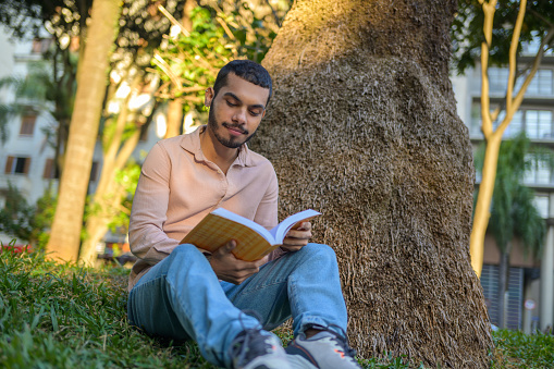 A man reading and enjoying the pages of the book sitting on a grass next to a tree