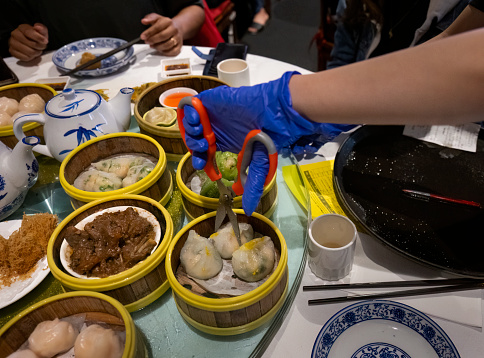 Woman hand with gloves holding scissors and cutting dumplings on a table full of Yumcha dishes. Pen and order paper on table.