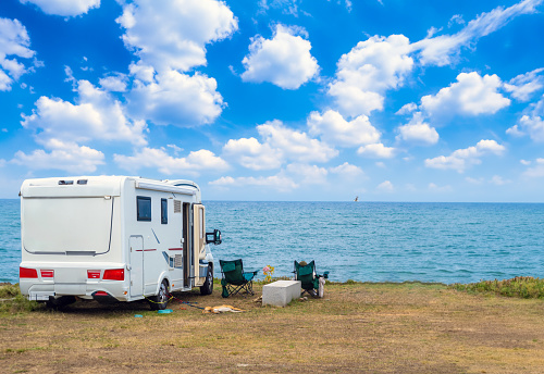 Camper van parked facing the sea from the top of a cliff under a blue sky with white clouds and two sun loungers overlooking the sea with a tourist lounging in the chair next to his dog on a leash.