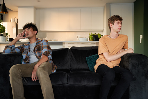 Young gay couple wearing casual clothing ignoring each other on sofa in living room at home
