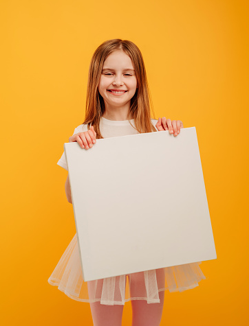 Cute pretty girl child holding white canvas and looking at the camera isolated on yellow background with copyspace. Portrait of smiling female kid holding linen with place for text