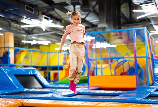 Pretty girl kid jumping on colorful trampoline at playground park and smiling. Caucasian preteen child during active entertaiments indoor